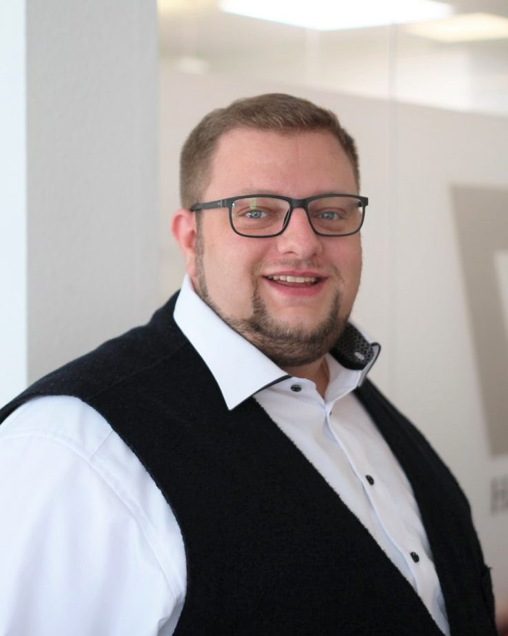 Mann mit Brille, hellem Hemd und dunkler Weste, lächelt freundlich in die Kamera.
